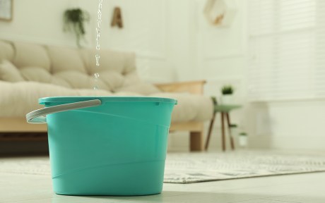 Decorative photo: Water dripping from ceiling into plastic bucket on floor in living room, space for text. Leaking roof