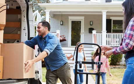 Photo of family moving into their first home; taking boxes out of a truck. Parents and two daughters.