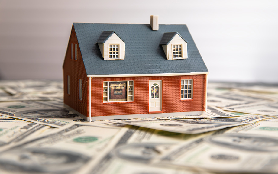 Decorative photo: play house with gray roof and red brick exterior, sitting on top of a spread out pile of $20 bills.