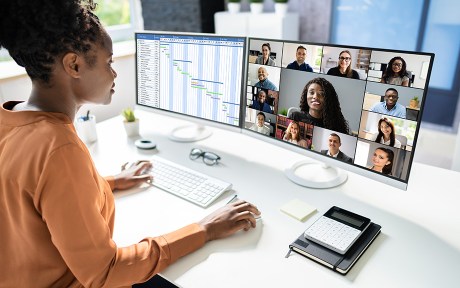 Photo of African American woman sitting at their desk looking at two screens; one screen has images of many people on a remote call, the other screen has a spreadsheet open.
