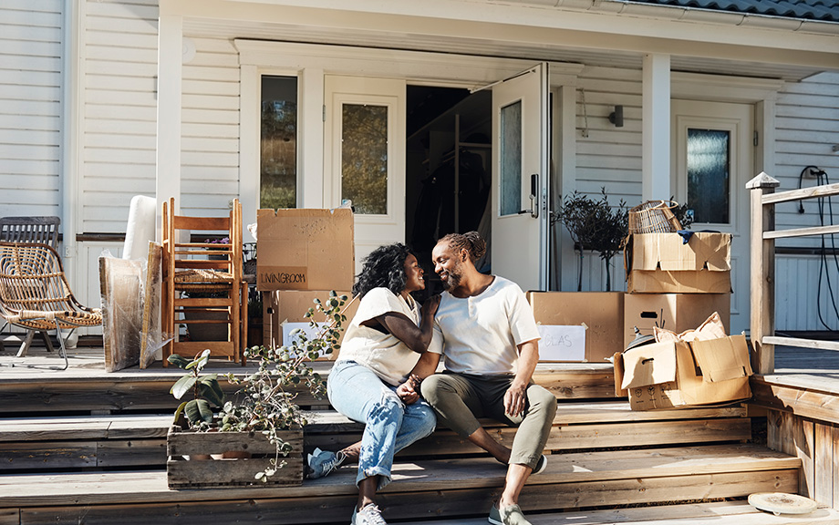 Young African American heterosexual couple sitting on the steps of a house with front door open and cardboard moving boxes around them.