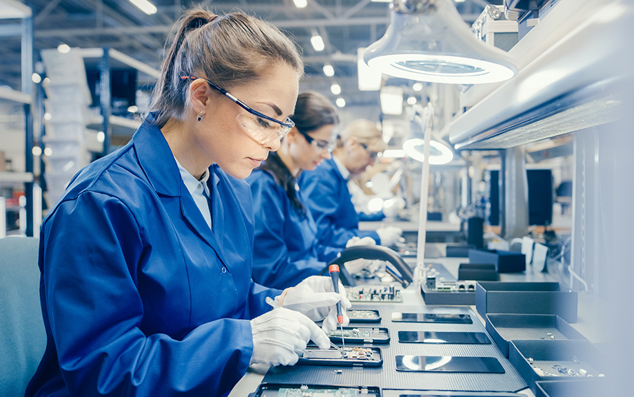 Photo of women in an electronics Factory Worker in Blue Work Coat and Protective Glasses is Assembling Smartphones with Screwdriver. High Tech Factory Facility with more Employees in the Background.