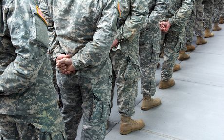 Photo: people in army fatigues lined up single file looking at their backs with the hands clasped behind them.