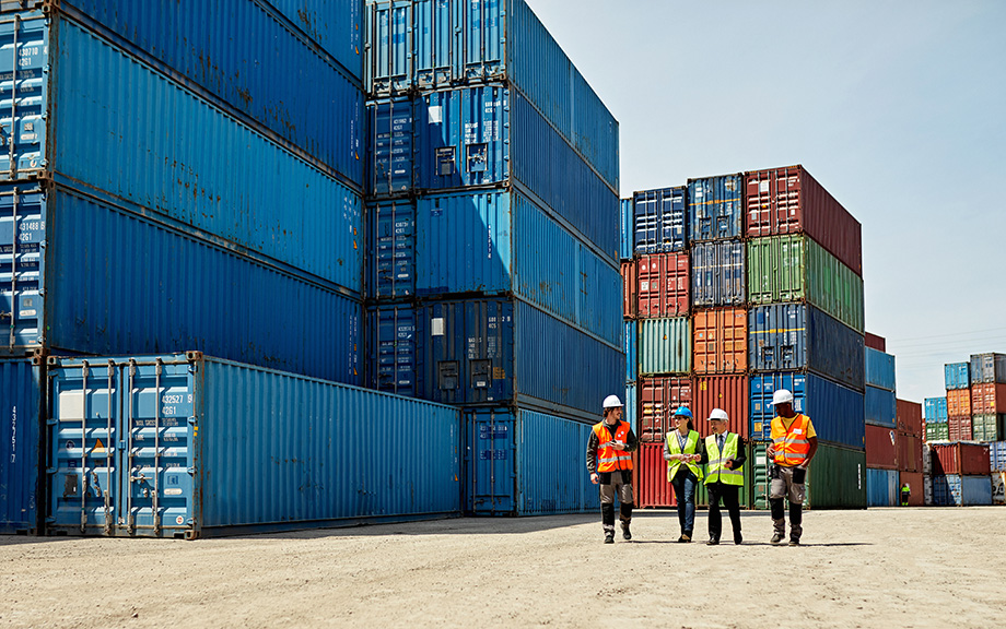 Photo: stacked shipping containers with a few worked in hard hats walking along side.