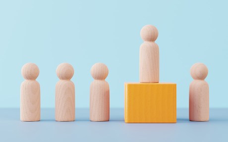 Photo: Wooden figurine pawn standing on wooden cube above other wooden figurines against minimal blue background. The concepts of competition, success, leadership, winning.