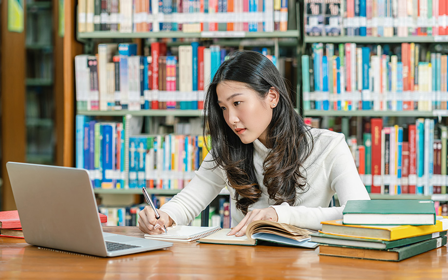 Photo of an Asian female college student in the library in front of shelves of books looking at her laptop and taking notes with books on the desk beside her.