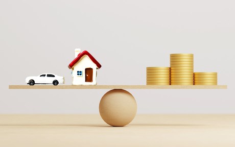 Coins stack on wooden seesaw on the right side, a car and house model on the left, equally balanced.