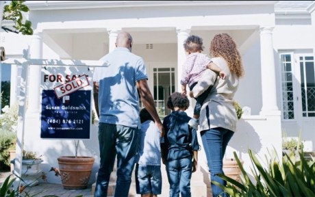 Decorative Image: A young family standing in front of the new home they purchased with the for sale/ sold sign next to them.