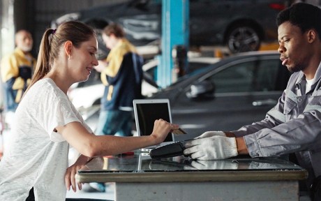 Photo of a car mechanic handing a woman customer a card reader in order to have her pay with credit card. She is placing her credit card on the reader.