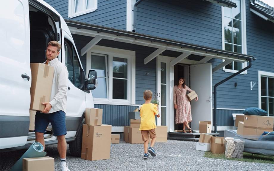 Photo: young couple with boy child bringing moving boxes into a newly purchased home that is blue clapboard and white trimmed windows. Boy is bringing his mother, who is standing in the doorway, a plant that looks like lavender.