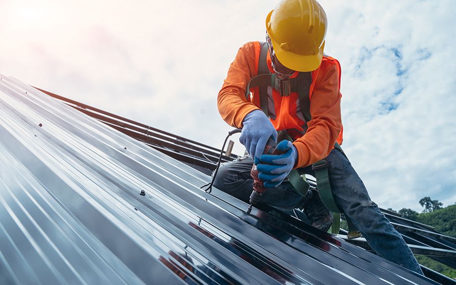Roofer worker in special protective work wear and gloves,using air or pneumatic nail gun and installing asphalt or bitumen shingle on top of the Metal Sheet roof.