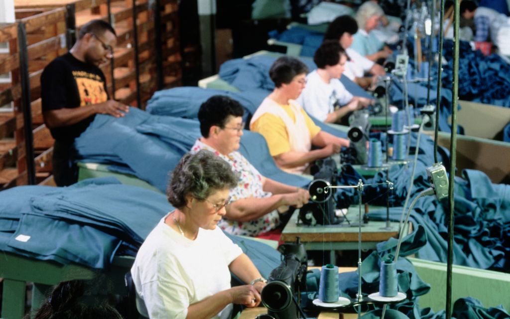 Photo of men and women sitting at sewing machines all in a row handling blue material.