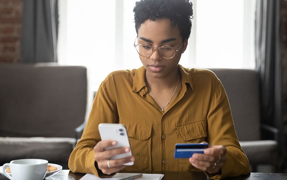 Serious young Black bank customer woman paying for purchase, domestic fees, bills by blue plastic credit card, using online payment app on smartphone, shopping on internet stores